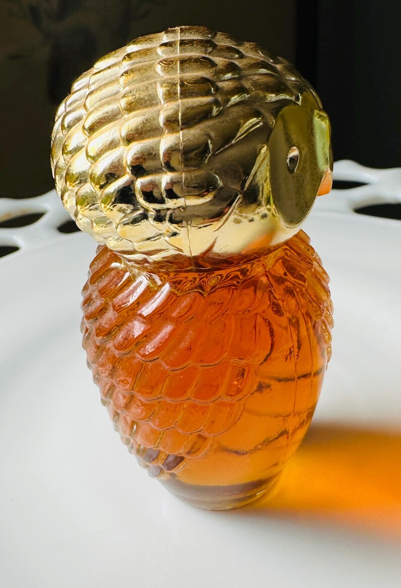 K&ouml;nnte beinhalten: Dekorative parf&uuml;mflasche in Eulenform. Der obere Teil ist goldfarben, der K&ouml;rper bernsteinfarben. Die Flasche hat eine strukturierte Oberfl&auml;che, die Federn &auml;hnelt. Augen und Schnabel der Eule sind sichtbar. Die Flasche steht auf einer wei&szlig;en Oberfl&auml;che.