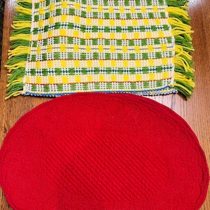 May include: A yellow, green, and white checkered placemat with fringe and a red oval placemat. The checkered placemat has a blue border. The red placemat has a quilted pattern.