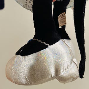 K&ouml;nnte beinhalten: Nahaufnahme von Fu&szlig; und Bein eines Pl&uuml;schtiers. Der Fu&szlig; ist ein gro&szlig;er, wei&szlig;er, glitzernder Schuh mit schwarzer Spitze und silberner Verzierung. Das Bein ist schwarz. Das Spielzeug ist wahrscheinlich eine Figur aus einem Zeichentrickfilm oder Film.