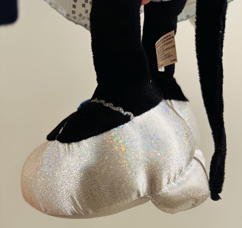 K&ouml;nnte beinhalten: Nahaufnahme von Fu&szlig; und Bein eines Pl&uuml;schtiers. Der Fu&szlig; ist ein gro&szlig;er, wei&szlig;er, glitzernder Schuh mit schwarzer Spitze und silberner Verzierung. Das Bein ist schwarz. Das Spielzeug ist wahrscheinlich eine Figur aus einem Zeichentrickfilm oder Film.