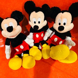 Puede incluir: Tres peluches de Mickey Mouse. Cada peluche tiene pelo negro, una cara bronceada, orejas negras y una nariz negra. Llevan pantalones cortos rojos con botones blancos, guantes blancos y zapatos amarillos. Los juguetes est&aacute;n colocados sobre una superficie naranja.