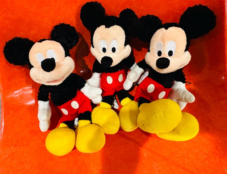 Puede incluir: Tres peluches de Mickey Mouse. Cada peluche tiene pelo negro, una cara bronceada, orejas negras y una nariz negra. Llevan pantalones cortos rojos con botones blancos, guantes blancos y zapatos amarillos. Los juguetes est&aacute;n colocados sobre una superficie naranja.