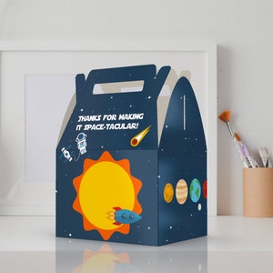 May include: A blue and orange party favor box with a space theme. The box features a sun, a rocket, a planet, and an astronaut. The text on the box reads "Thanks for making it space-tacular!"