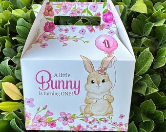 May include: A white favor box with a pink bunny rabbit illustration and the text "A little Bunny is turning ONE!"  The box has a pink balloon with the number 1 on it. The box is decorated with pink flowers.
