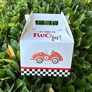 May include: White favor box with a red race car and black and white checkered pattern. The box has a handle and says "Growing Up Two Fast" in red and black text.