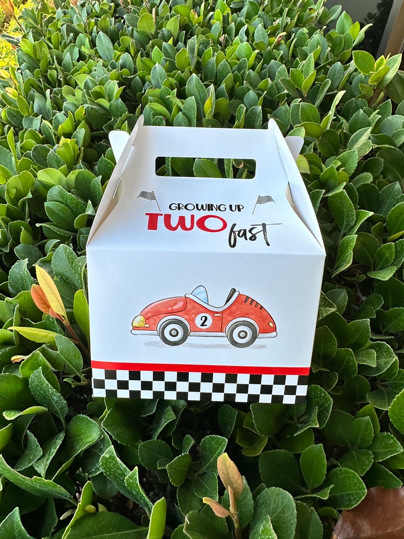 May include: White favor box with a red race car and black and white checkered pattern. The box has a handle and says "Growing Up Two Fast" in red and black text.