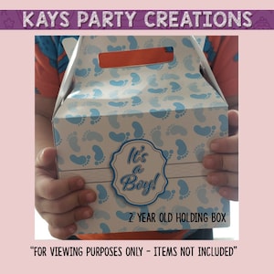 Op de afbeelding: Een blauwe en witte cadeaudoos met een blauw voetstappenpatroon. De doos heeft een handvat en een label met de tekst "It's a Boy!" en "2 Year Old Holding Box". De tekst "ALLEEN VOOR BEKIJKDOELEINDEN - ARTIKELEN ZIJN NIET INBEGREPEN" staat onder de doos.