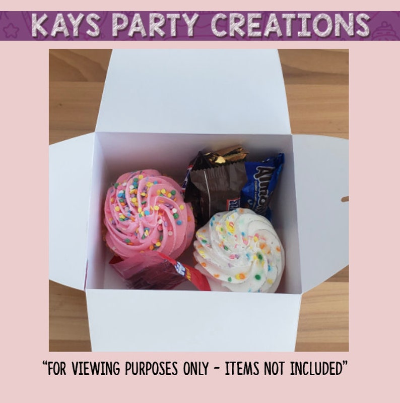 Op de afbeelding: Een witte doos met een roze en witte cupcake, een witte cupcake met sprinkles en een chocolade reep erin. De doos is open en de items zijn zichtbaar. "Alleen voor kijkdoeleinden - items niet inbegrepen" staat onderaan de afbeelding.