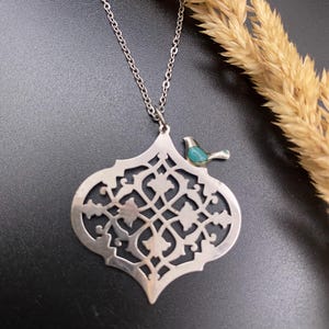 May include: A silver-toned pendant necklace featuring an openwork design with a small turquoise bird accent. The pendant has an intricate, symmetrical pattern. The necklace is displayed on a dark surface with dried wheat stalks.