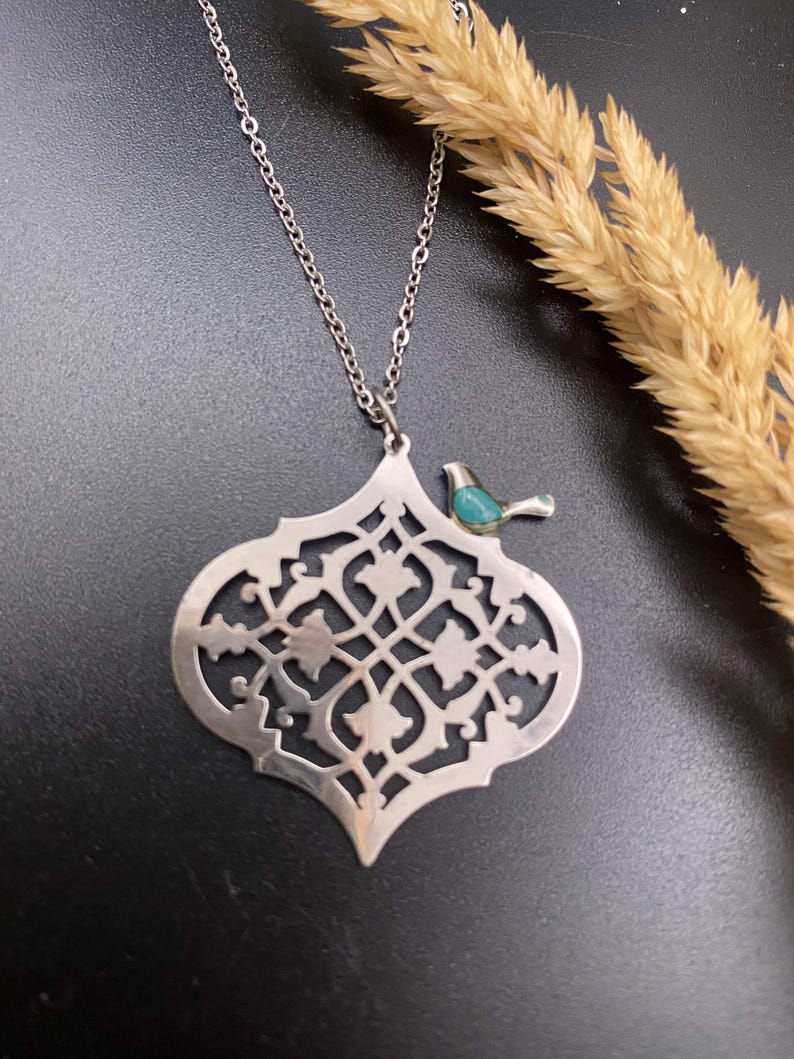 May include: A silver-toned pendant necklace featuring an openwork design with a small turquoise bird accent. The pendant has an intricate, symmetrical pattern. The necklace is displayed on a dark surface with dried wheat stalks.