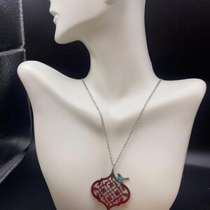 May include: A silver necklace with a red filigree pendant and a small turquoise bird accent. The pendant has an intricate openwork design. The necklace is displayed on a white mannequin bust against a dark background.