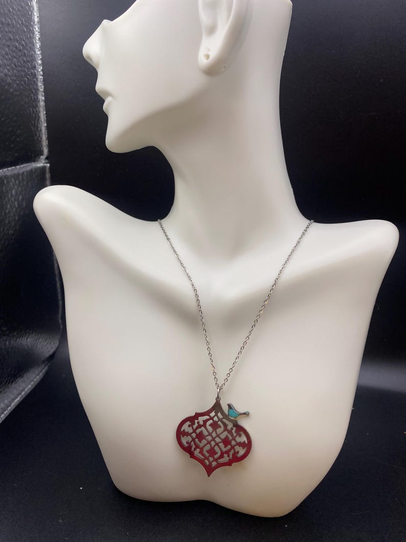 May include: A silver necklace with a red filigree pendant and a small turquoise bird accent. The pendant has an intricate openwork design. The necklace is displayed on a white mannequin bust against a dark background.