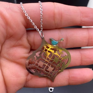 May include: Silver-tone necklace with a pendant featuring an openwork design and a small turquoise bird. The pendant has a gold-colored interior and a decorative pattern. The necklace is displayed against a dark background.
