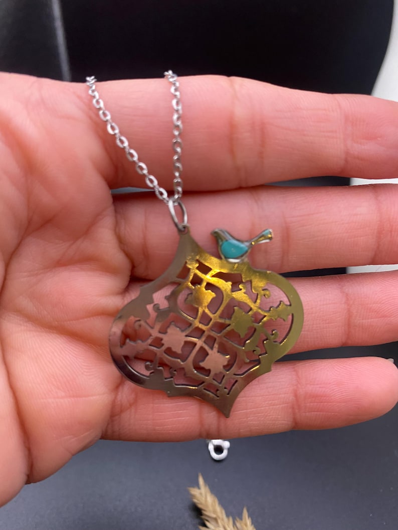 May include: Silver-tone necklace with a pendant featuring an openwork design and a small turquoise bird. The pendant has a gold-colored interior and a decorative pattern. The necklace is displayed against a dark background.