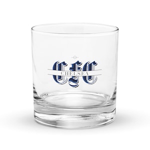 May include: A clear, round rocks glass with the text "CFC CHELSEA" in blue and white. The glass has a thick base and is designed for serving spirits or cocktails. The text is centered on the glass.