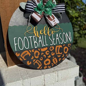 K&ouml;nnte beinhalten: Rundes Holzschild mit Fu&szlig;ball-Motiv. Das Schild zeigt den Text "hello FOOTBALL SEASON!" in Gelb und Wei&szlig;. Es ist mit Fu&szlig;b&auml;llen und einer Schleife mit Fu&szlig;ball- und Feldmustern verziert.