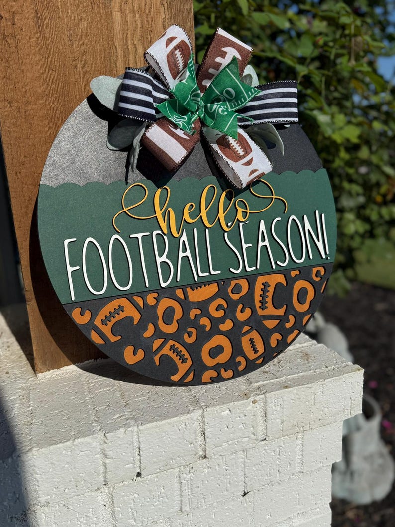 K&ouml;nnte beinhalten: Rundes Holzschild mit Fu&szlig;ball-Motiv. Das Schild zeigt den Text "hello FOOTBALL SEASON!" in Gelb und Wei&szlig;. Es ist mit Fu&szlig;b&auml;llen und einer Schleife mit Fu&szlig;ball- und Feldmustern verziert.