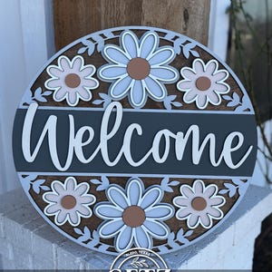 May include: A round wooden sign with the word "Welcome" in white script. The sign features a floral design with blue, brown, and white daisies. The sign is layered and has a dark gray background.