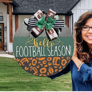 K&ouml;nnte beinhalten: Runder Holzschild mit gr&uuml;nem und schwarzem Hintergrund, auf dem der Text "hello FOOTBALL SEASON!" und Football-Muster zu sehen sind. Das Schild ist mit einer Schleife und Football-Akzenten verziert, perfekt f&uuml;r die T&uuml;rdekoration.