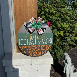 K&ouml;nnte beinhalten: Runder, dekorativer Holzschild mit dem Text "hello FOOTBALL SEASON!" in wei&szlig;er und goldener Schrift. Das Schild hat ein gr&uuml;nes und orangefarbenes Football-Design und ist mit einer dekorativen Schleife verziert.