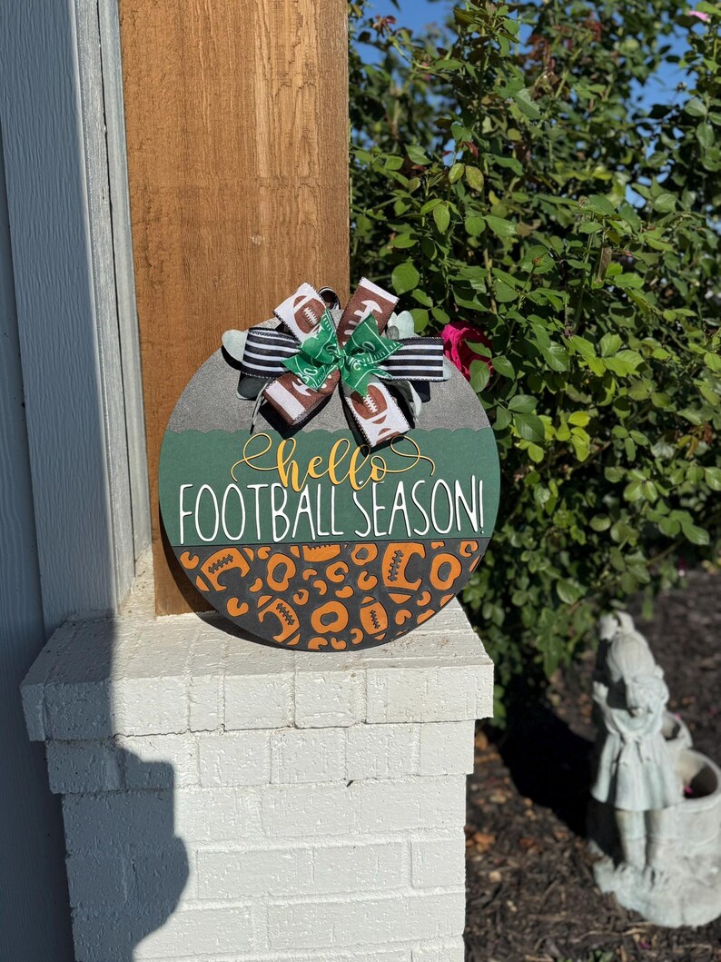 K&ouml;nnte beinhalten: Runder, dekorativer Holzschild mit dem Text "hello FOOTBALL SEASON!" in wei&szlig;er und goldener Schrift. Das Schild hat ein gr&uuml;nes und orangefarbenes Football-Design und ist mit einer dekorativen Schleife verziert.
