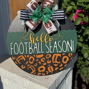 K&ouml;nnte beinhalten: Runder Holzschild mit Fu&szlig;ball-Motiv. Das Schild hat einen gr&uuml;nen und schwarzen Hintergrund mit den Worten "Hello Football Season!" in Wei&szlig; und Gelb. Eine dekorative Schleife mit Fu&szlig;ball- und Feldmustern ziert die Oberseite.