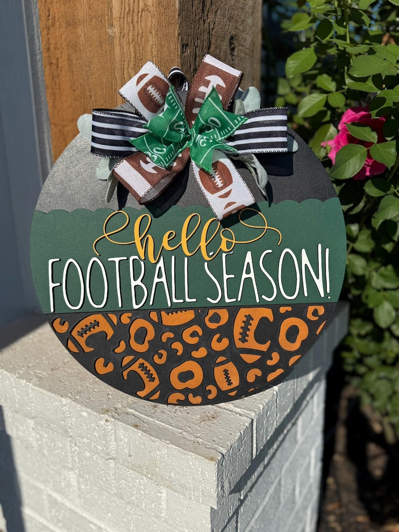 K&ouml;nnte beinhalten: Runder Holzschild mit Fu&szlig;ball-Motiv. Das Schild hat einen gr&uuml;nen und schwarzen Hintergrund mit den Worten "Hello Football Season!" in Wei&szlig; und Gelb. Eine dekorative Schleife mit Fu&szlig;ball- und Feldmustern ziert die Oberseite.