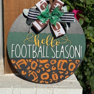 K&ouml;nnte beinhalten: Runder Holzschild mit gr&uuml;nem und grauem Hintergrund, mit dem Text "hello FOOTBALL SEASON!". Das Schild ist mit einer Schleife aus Football-Themenband und einem Leopardenmuster mit Fu&szlig;b&auml;llen verziert.