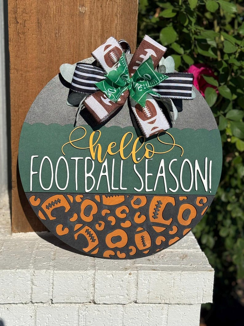 K&ouml;nnte beinhalten: Runder Holzschild mit gr&uuml;nem und grauem Hintergrund, mit dem Text "hello FOOTBALL SEASON!". Das Schild ist mit einer Schleife aus Football-Themenband und einem Leopardenmuster mit Fu&szlig;b&auml;llen verziert.
