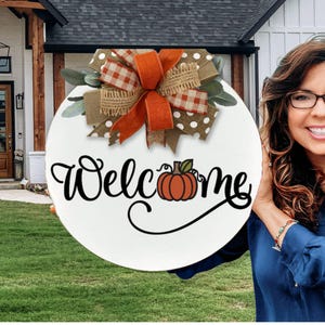 May include: A round white wooden welcome sign with a pumpkin illustration and the word "Welcome" in black script. The sign is decorated with a bow made of orange, brown, and polka dot ribbons and greenery.