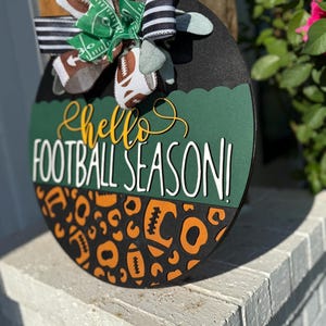 K&ouml;nnte beinhalten: Runder, schwarzer Holzschild mit dem Text "Hello Football Season!" in Wei&szlig; und Gelb. Das Schild hat einen gr&uuml;nen und schwarzen, gewellten Rand und ein Leopardenmuster mit orangefarbenen Fu&szlig;b&auml;llen. Eine dekorative Schleife mit Fu&szlig;ball- und Streifenb&auml;ndern ziert das Schild.