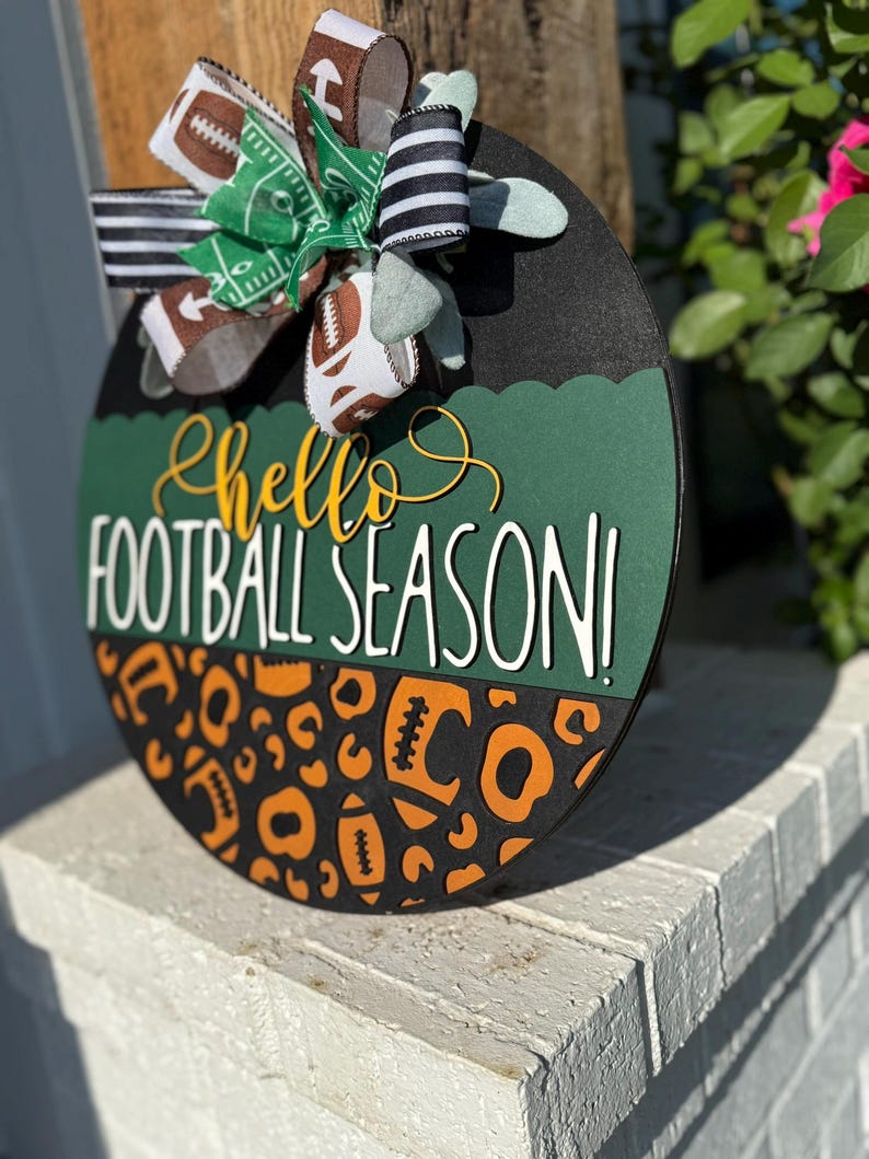 K&ouml;nnte beinhalten: Runder, schwarzer Holzschild mit dem Text "Hello Football Season!" in Wei&szlig; und Gelb. Das Schild hat einen gr&uuml;nen und schwarzen, gewellten Rand und ein Leopardenmuster mit orangefarbenen Fu&szlig;b&auml;llen. Eine dekorative Schleife mit Fu&szlig;ball- und Streifenb&auml;ndern ziert das Schild.