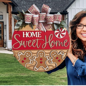 May include: A round wooden sign with the words "Home Sweet Home" in white and gold lettering. The sign features gingerbread men and a peppermint candy design, with a bow on top. The sign is a festive decoration.