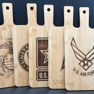 May include: A collection of bamboo cutting boards with handles, each engraved with a different U.S. military emblem. The boards feature the emblems of the Marine Corps, Army, and Air Force, along with the Space Force logo. The boards are light brown.
