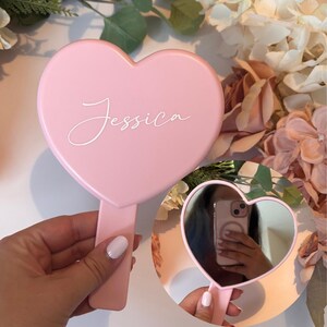 Pode incluir: Um espelho de mão rosa em forma de coração com o nome "Jessica" em escrita branca. O espelho tem uma alça e uma superfície refletora. A imagem mostra um close-up do espelho e seu reflexo.