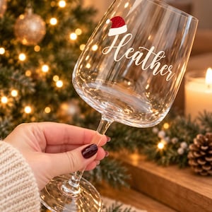 Puede incluir: Una copa de vino de cristal transparente con el nombre Heather en letra cursiva blanca, decorada con un pequeño gráfico de gorro de Papá Noel rojo y blanco brillante sobre la letra H, sobre un fondo festivo.