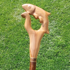 Op de afbeelding: Houten wandelstok met een uitgesneden visontwerp op het handvat. De vis is lichtbruin en de stok heeft een donkerbruin onderste gedeelte met een messingkleurige band. De achtergrond is groen gras.