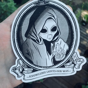 Könnte beinhalten: Schwarzweiß-Illustration eines Außerirdischen, der eine Kapuzenrobe trägt und Kristalle hält. Das Bild ist in einem Oval gerahmt mit dem Text "...KNOWLEDGE LIGHTS OUR WAY..." unten.