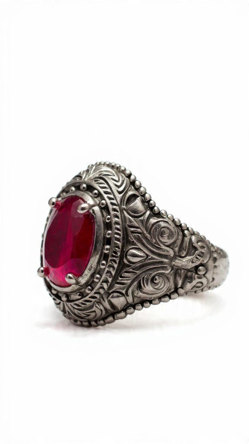 May include: A silver ring with an oval, deep red gemstone. The ring has detailed scrollwork and bead accents around the stone and band. The ring is set against a white background. The ring is an antique style.