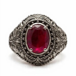 May include: A silver ring with a large, oval, deep red gemstone. The ring has detailed, ornate patterns around the gemstone and on the band, giving it a vintage look. The ring is set against a white backdrop, highlighting the design.