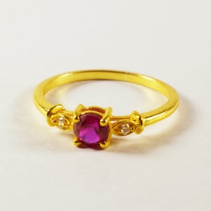 May include: A gold ring featuring a single red gemstone and two smaller clear gemstones. The ring is displayed against a white backdrop.