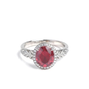May include: A silver ring featuring a large, oval-cut, deep red gemstone surrounded by a halo of smaller, clear stones. The band has an engraved leaf design. A classic and elegant piece of jewelry.