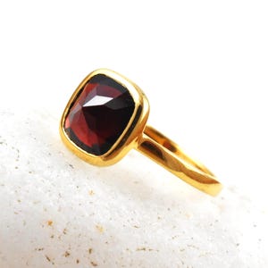 May include: A gold ring featuring a cushion-cut, deep red gemstone. The gemstone is set in a gold bezel, and the ring's band is also gold. The ring is displayed on a white, textured surface. This piece is a jewelry item.