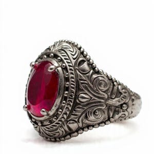 May include: A silver ring with an oval, deep red gemstone. The ring has detailed carvings and a beaded border. The ring is set against a white background. The design is antique-inspired.