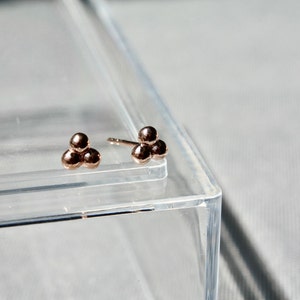 Könnte beinhalten: Ohrstecker aus Roségold. Jeder Ohrring hat drei kleine, runde Perlen, die in einer dreieckigen Anordnung angeordnet sind. Die Ohrringe werden auf einer transparenten Oberfläche mit einem weichen, neutralen Hintergrund präsentiert.