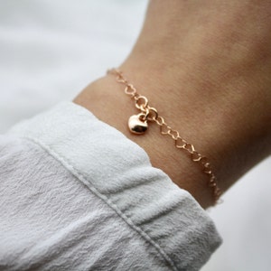 Könnte beinhalten: Ein zartes Armband aus Roségold mit einer Kette aus kleinen herzförmigen Gliedern. Ein kleiner Herzanhänger baumelt am Armband. Das Armband wird an einem Handgelenk vor weißem Hintergrund gezeigt.