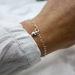 Könnte beinhalten: Ein filigranes Silberarmband mit einer Kette aus kleinen herzförmigen Gliedern. Ein größerer Herzanhänger baumelt von der Mitte. Das Armband wird an einem Handgelenk getragen, vor einem weißen Stoffhintergrund.