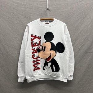 May include: A white long-sleeve crewneck sweatshirt featuring a large graphic of Mickey Mouse on the front. The character is depicted in black, white, and tan, with the word MICKEY printed vertically in bold red letters along the left side.