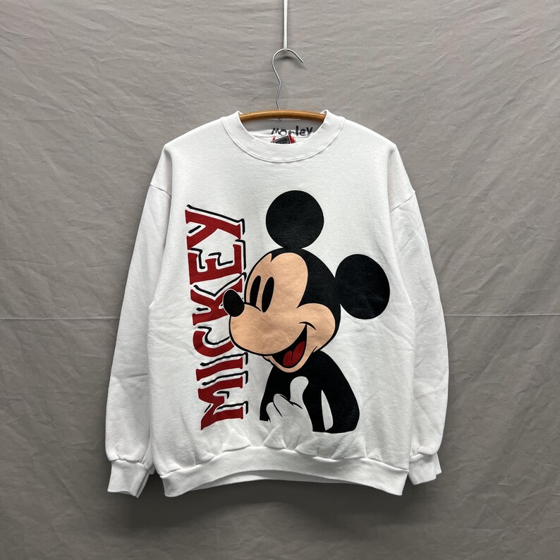May include: A white long-sleeve crewneck sweatshirt featuring a large graphic of Mickey Mouse on the front. The character is depicted in black, white, and tan, with the word MICKEY printed vertically in bold red letters along the left side.