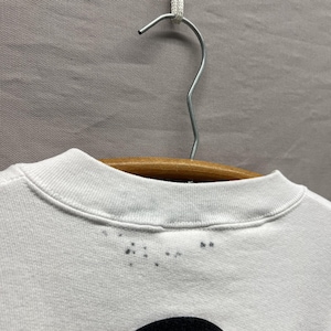 May include: A white crew neck t-shirt hanging on a wooden hanger. The collar features a ribbed neckline with a series of small, dark grey spots or stains scattered along the front edge just below the seam of the collar, visible against the fabric.